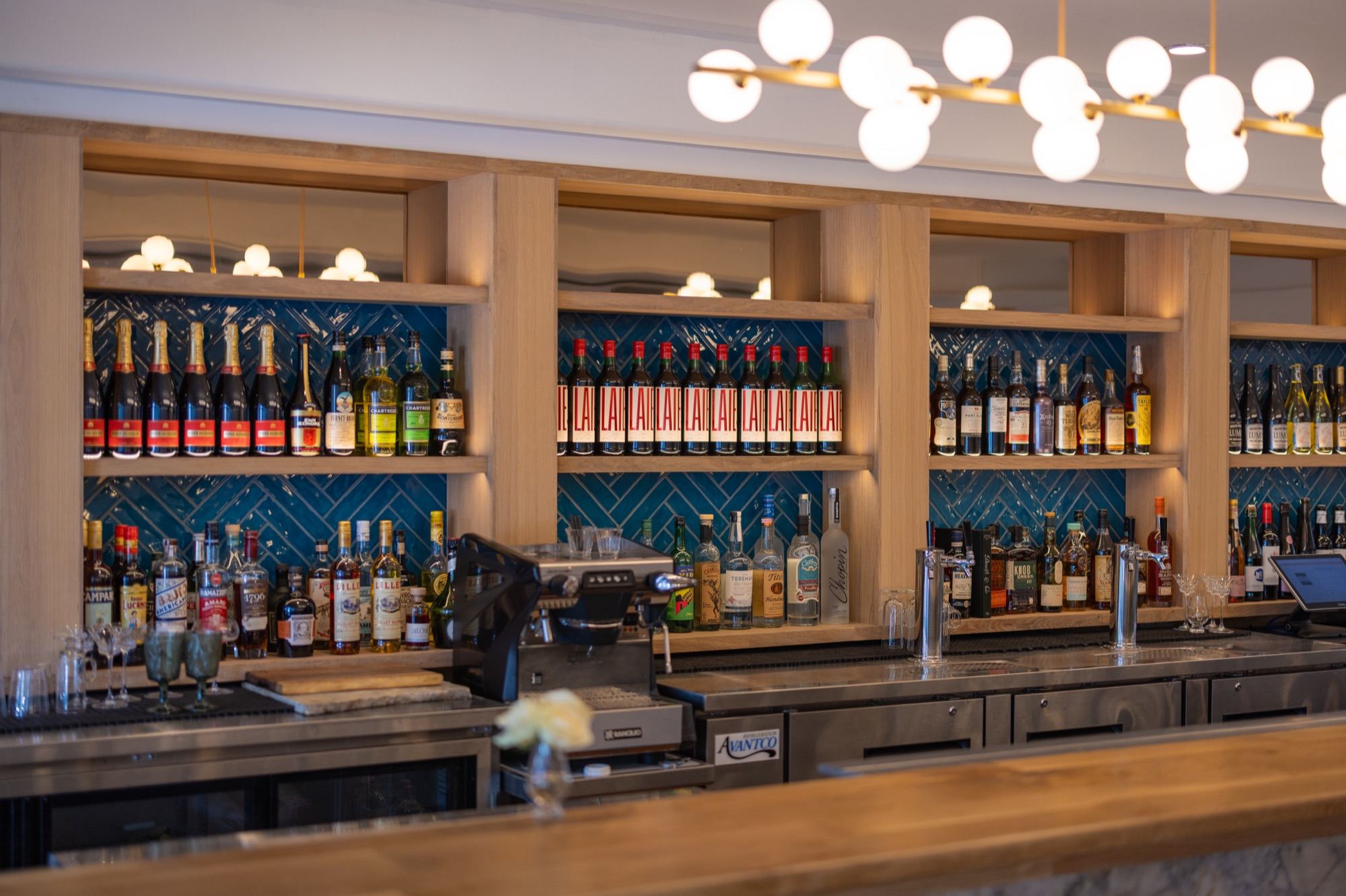 Wooden back-bar shelves on blue herringbone tile holding rows of bottles, with a globe chandelier above the bar.
