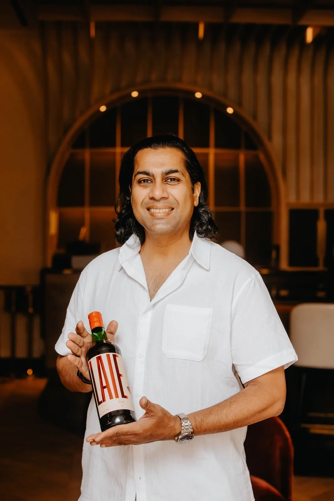 Raj Nallapothola in a white shirt, smiling and holding a bottle of LAIE Rojo in a warmly lit restaurant.
