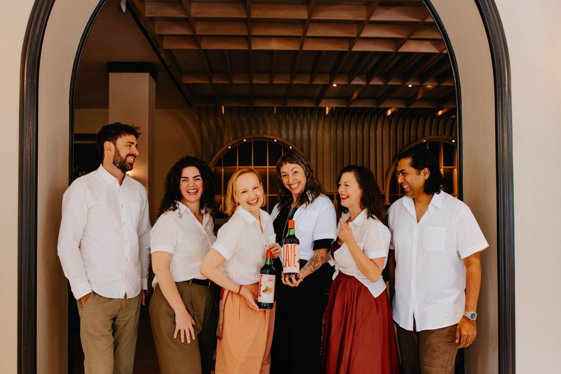 The LAIE team, six people in white shirts, laughing together in an arched doorway, two of them holding bottles of LAIE Rojo.