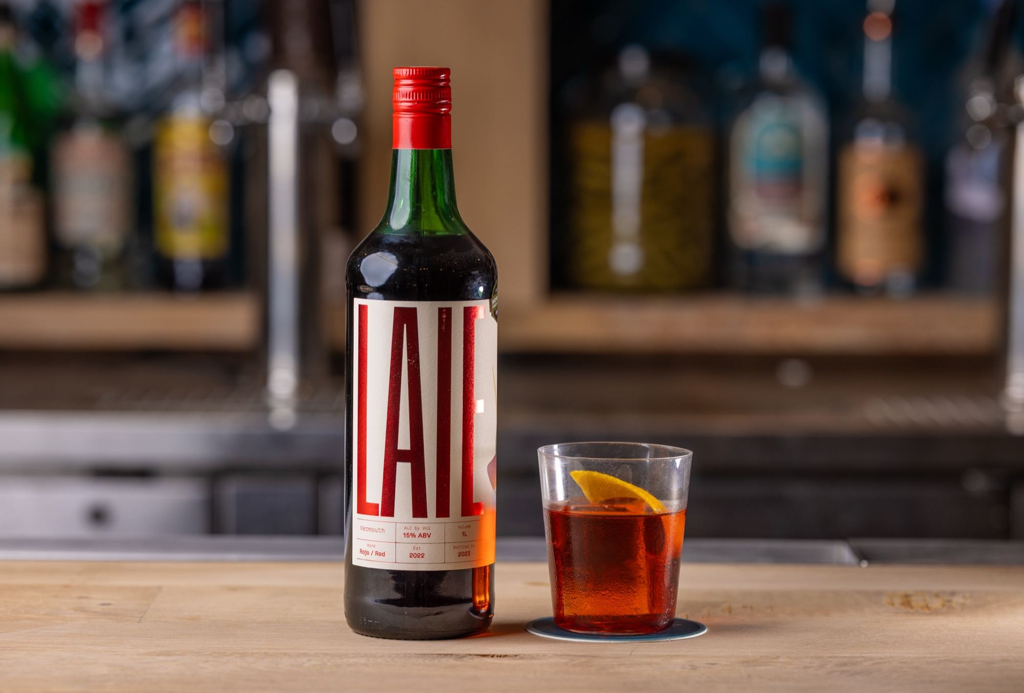 A LAIE Rojo Vermouth bottle beside a short glass of red-amber drink with an orange slice on a wooden bar.