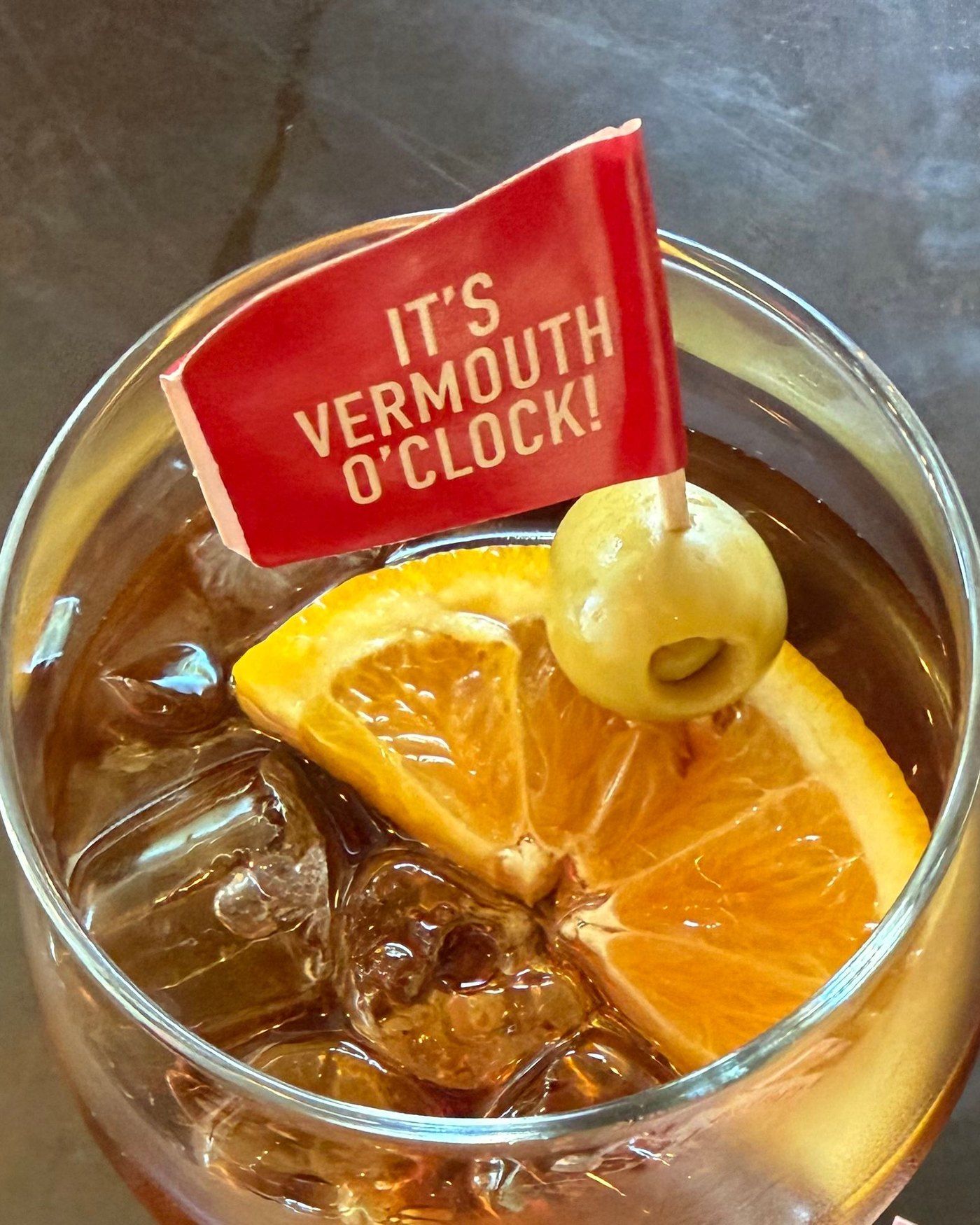 Overhead view of a glass of LAIE Rojo over ice with an orange slice and a green olive on a red flag pick.