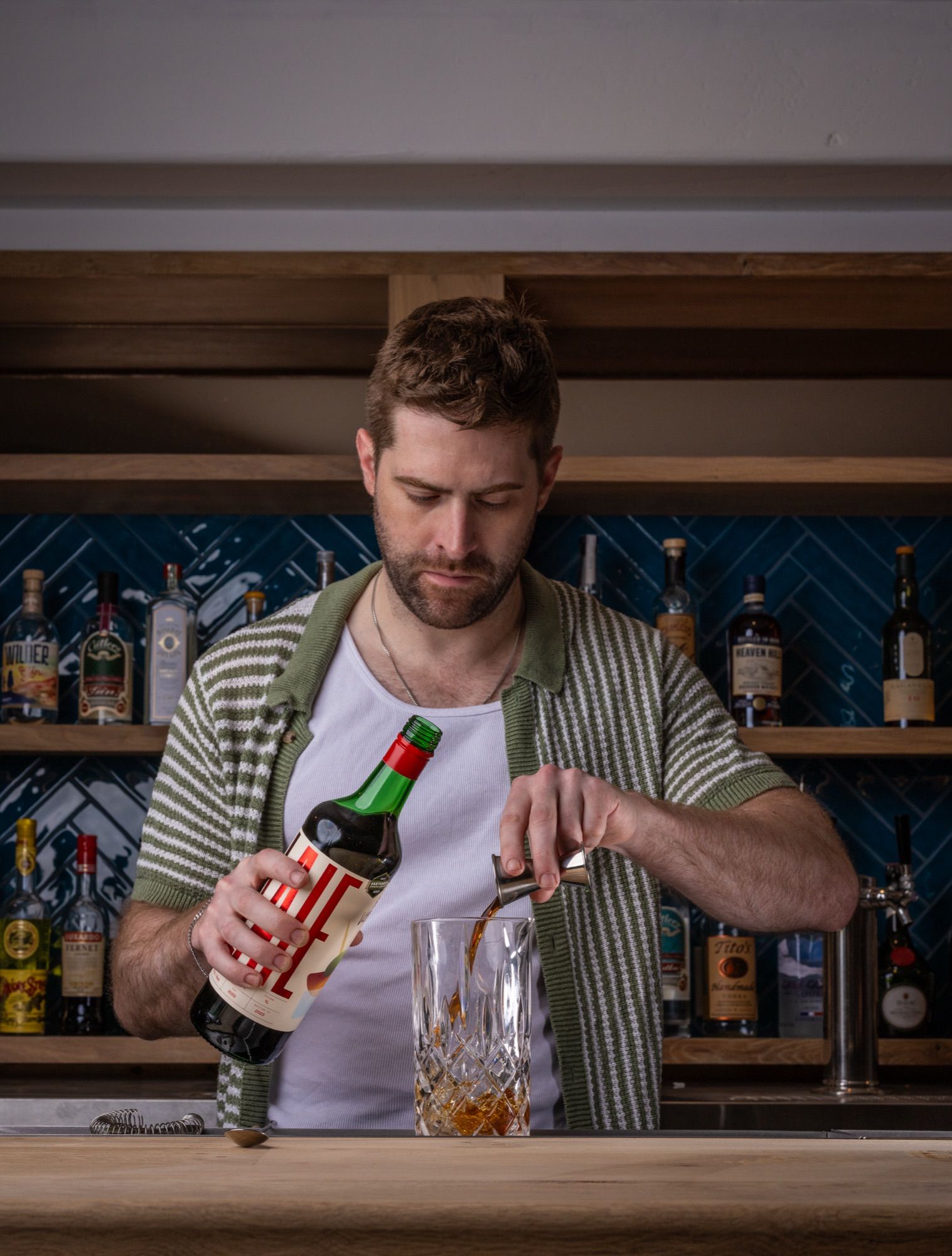 A bartender in a striped cardigan pours LAIE Rojo Vermouth from a jigger into a crystal mixing glass in front of a lit back bar.